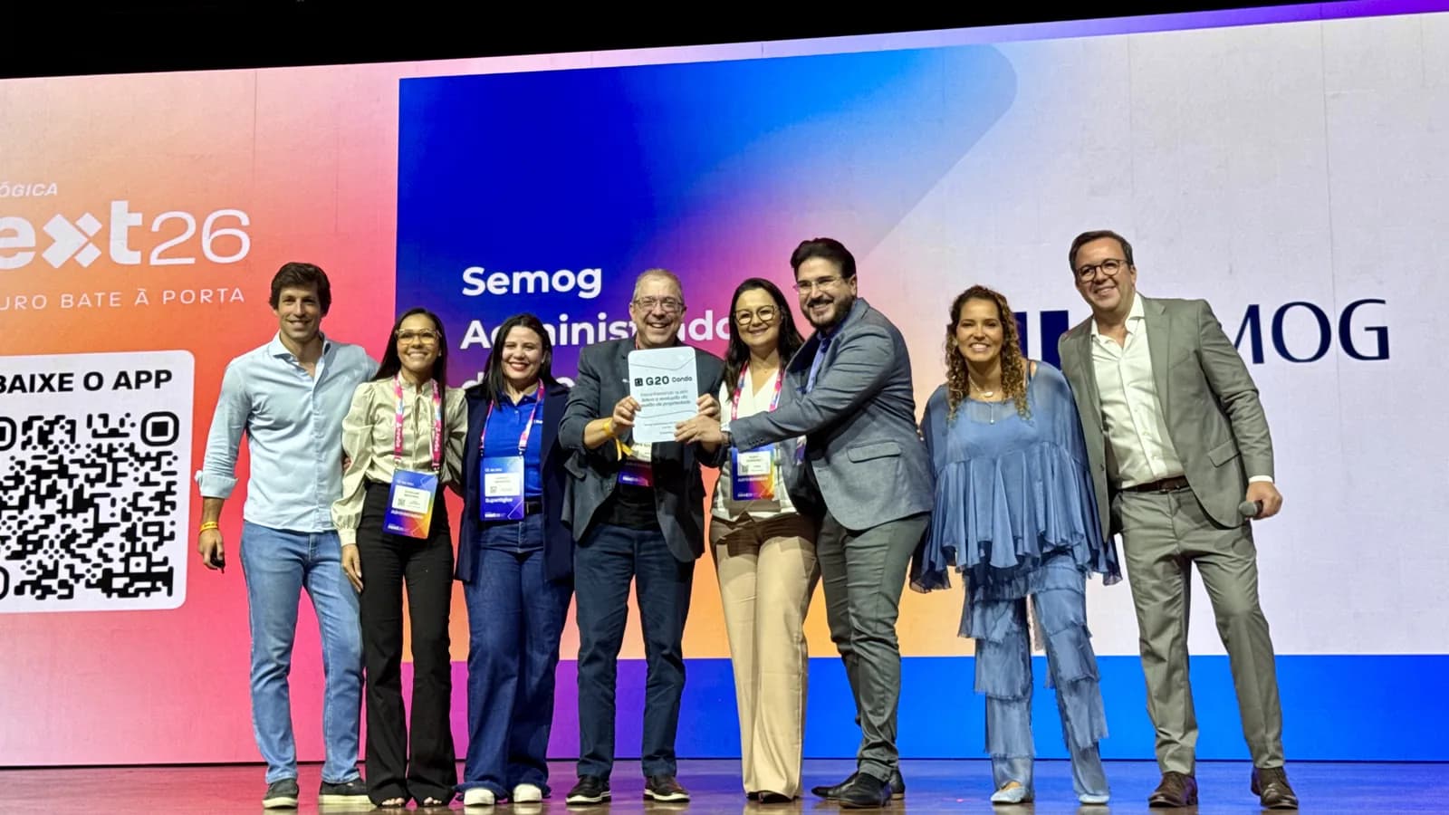 Equipe da Semog no palco do Superlógica Next 2026 segurando a placa do G20 Condo, com o logotipo da empresa no telão